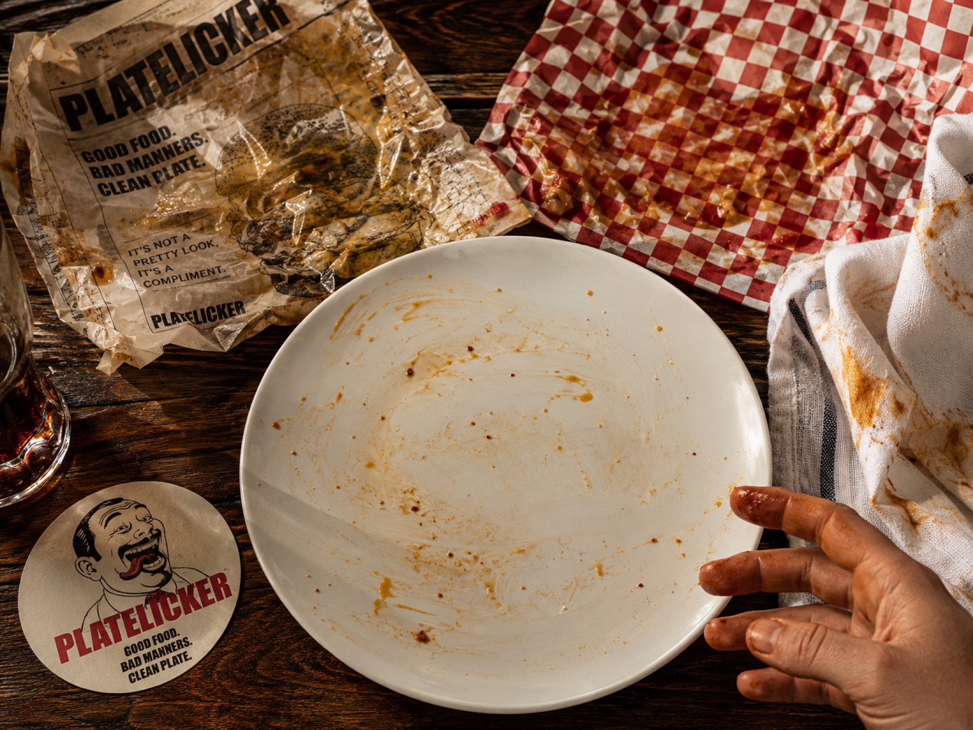 A plate wiped clean of curry, a sauce-stained hand beside it, a greasy PlateLicker wrapper on a dark wooden table